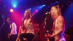 Der Sänger von Towers Of London steht bei Bootleg im April 2006 in der Mitte der Bühne, zwischen den Gitarristen und zeigt mit dem Finger nach links.