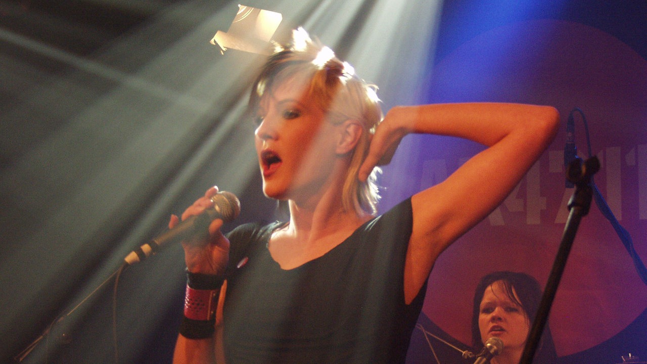 Die Frontfrau von AK4711 steht bei Bootleg im April 2006 im Scheinwerferlicht und greift sich an den Hinterkopf.