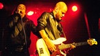 Ein kurzrasierter Bassist spielt, daneben der Sänger.