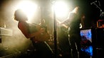 Der Gitarrist und der Bassist der Band stehen beide vor gleizend weißem Licht in Dunst der Bühne und spielen auf ihren Instrumenten.