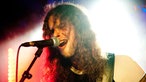 Der blau-rot angestrehlte Bassist hat die Haare ins Gesicht hängen und singt mit weit offenem Mund in das Mikrofon.