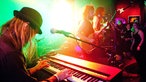 Im Vordergrund ist der Keyboarder zu sehen welcher mit nach unten geneigtem Kopf spielt, während im Hintergrund der Rest der Band in grünem Licht erstrahlt.