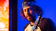 Einer der Gitarristen hat ein dunkles Bandana um seine Stirn gewickelt und sieht angestrengt nach unten auf seine Gitarre.