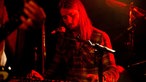 Der Keyboarder der Band sieht etwas gelangweilt nach unten auf sein Instrument und wird dabei rot von der Seite angestrahlt.