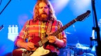 Der rundliche Frontmann der Band sieht angestrengt nach unten und spielt auf seiner Gitarre.