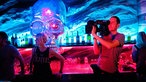 Ein Kameramann steht mit voller Ausrüstung an der Bar und ist am Filmen. Im Hintergrund sieht man einen riesigen gläsernen Schädel an der Wan hängen dessen eines Auge rot erleuchtet ist.