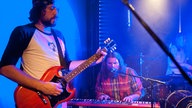 Ein weiters Bandmitglied steht mit einer Gitarre im Vordergrund währen im Hintergrund der Keyboarder am Mikrofon mitsingt.