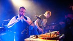 Zwei Musiker von Antlered Man, der eine spielt eine kleine Flöte, der andere Bass. Vor ihnen ein Tisch mit diversen Effektgeräten.