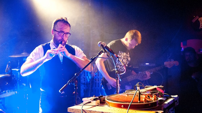 Zwei Musiker von Antlered Man, der eine spielt eine kleine Flöte, der andere Bass. Vor ihnen ein Tisch mit diversen Effektgeräten.