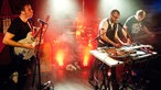 Aktionsreiche Aufnahme aller Antlered Man Musiker auf der Bühne: hinten der Drummer, davor Bassist, Gitarrist und der Effekte spielende Multiinstrumentalist - alle in Aktion.