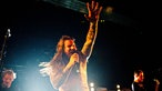 Der Sänger singt mit zugekniffenen Augen, die Haare fliegen, eine hand reckt er beschwöhrend in die Höhe.
