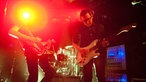 Im Hintergrund der Schlagzeuger, davor Bassist und Gitarrist der Band. Ausserdem ist ein Geräte-Rack zu sehen, an dem die vielen Kabel der Instrumente angeschlossen sind.