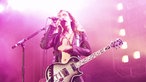 Mann mit langen Haaren, Lederjacke und nacktem Bauch singt im pinken Scheinwerferlicht.