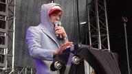Winson mit Kaputzenpullover und zwei Mikrofonen in der Hand steht an einem Keyboard.