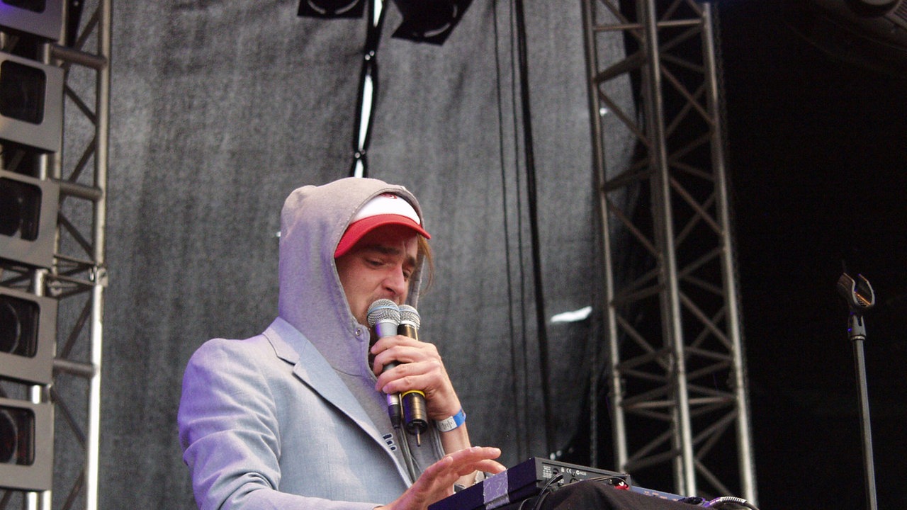 Winson mit Kaputzenpullover und zwei Mikrofonen in der Hand steht an einem Keyboard.