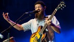 Harrison Stafford singt mit umgehängter Gitarre und gehobener Hand.