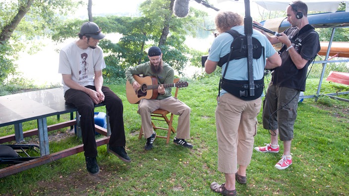 Groundation unplugged auf einer Wiese auf Holzstühlen während ein Kamerateam aufnimmt