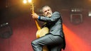 Der Gitarrist trägt eine schwarze Anzughose, Hemd und Sakko. Er spielt Akkustik-Gitarre