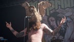 Der Sänger von Kvelertak, mit tätowiertem, nacktem Oberkörper. Er hat sich eine ausgestopfte Eule wie eine Maske über das Gesicht gestülpt und singt durch das Gefieder hindurch in das Mikrofon.