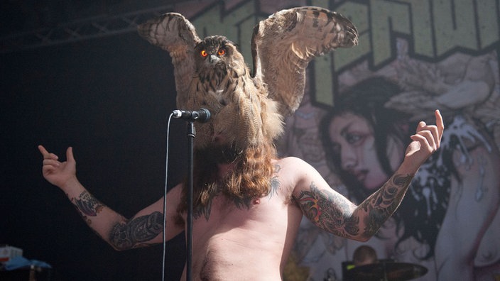 Der Sänger von Kvelertak, mit tätowiertem, nacktem Oberkörper. Er hat sich eine ausgestopfte Eule wie eine Maske über das Gesicht gestülpt und singt durch das Gefieder hindurch in das Mikrofon.