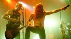 Der Sänger richtet das Mikrofon gen Publikum, während der schnurrbärtige Bassist spielt und singt.