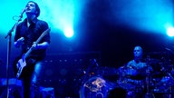Gitarrist spielt Gitarre und singt in ein Mikrofon, schräg links von Ihm spielt ein Mann das Schlagzeug, im Hintergrund sieht man blau strahlende Scheinwerfer