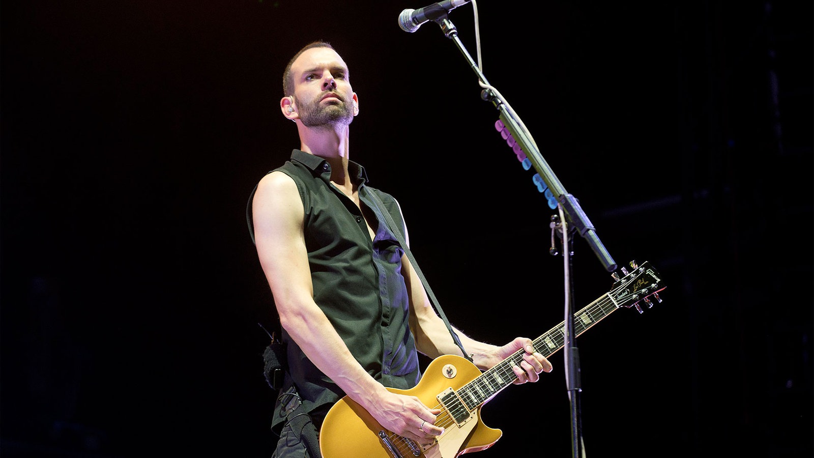 Gitarrist mit schwarzem Shirt, kurzem Haar und 3-Tage-Bart spielt eine gelbliche Gitarre; im Vordergrund steht ein Mikrofon