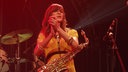 The Zutons: Eine Frau im gelben Kleid hat ein Saxophon umgehängt und klatscht im roten Bühnenlicht