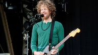 Gitarrist von Dispatch mit wilden Bartwuchs und einer Lockenfrisur singt energievoll ins Mikrofon