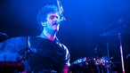Der Drummer der Band singt neben dem Schlagzeug spielen auch an einem kleinen Mikrofon mit, dass von oben herunter hängt.