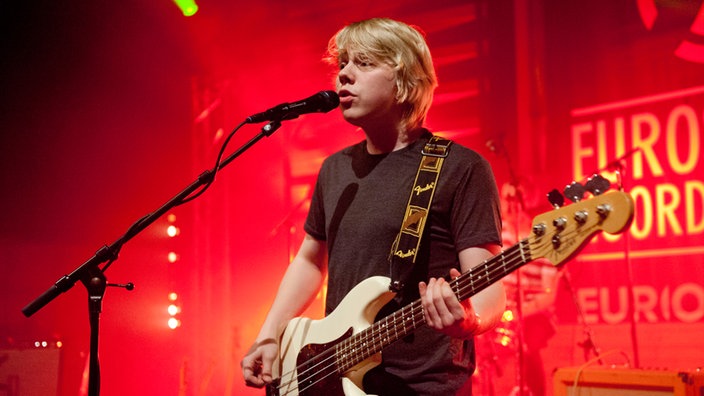Blonder junger Mann mit Gitarre singt in ein Mikrofon im roten Bühnenlicht.