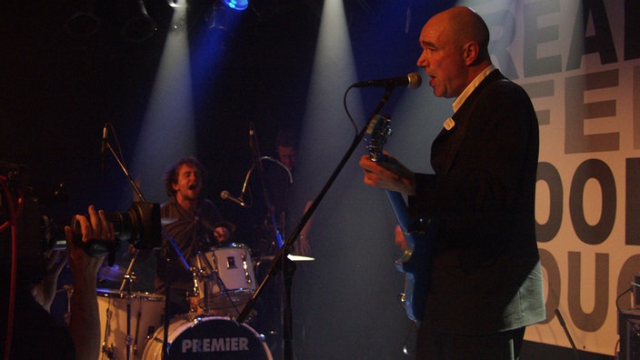 John Watts singt mit Gitarre und Schlagzeug im Hintergrund bei Bootleg im Oktober 2005