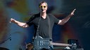 Mann mit gelber Sonnenbrille, umgehängter Gitarre und schwarzem T-Shirt breitet seine Arme aus.