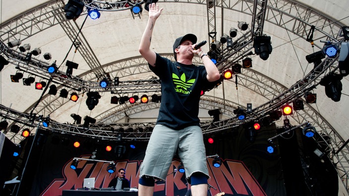 Ein Rapper mit kurzen Hosen steht auf der Bühne und hebt die Hand