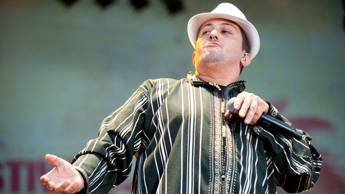 Frontmann Hakim Meridja posiert mit weissem Hut und Mikrofon auf der Bühne des Summerjam. Er streckt die rechte Hand aus und zeigt einen einstudierten, arroganten Blick 