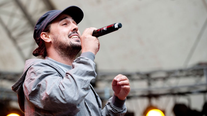 Die österreichische Band Iriepathie steht auf der Bühne. Frontmann "Professa" trägt eine graue Jacke und hält in der echten Hand das Mikrofon.