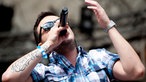 Backing-Vocalist „Syrix“ trägt ein karriertes Hemd und Sonnenbrille. Er singt in sein Funkmikrofon während er die linke Hand hebt