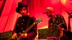 Ein Gitarrist trägt Hut und einen Frack mit Manschettenknöpfen. Im Hintergrund spielt der Keyboarder der Band
