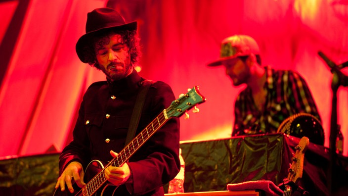 Ein Gitarrist trägt Hut und einen Frack mit Manschettenknöpfen. Im Hintergrund spielt der Keyboarder der Band