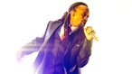"Madcon" Sänger  Tshawe Baqwa steht in einem gleisend hellen Licht und performed einen Song. Er trägt einen Anzug darunter ein weisses Hemd und eine rote Krawatte. Das ihn völlig umgebende weisse Licht lässt ihn wie ein Engel erscheinen