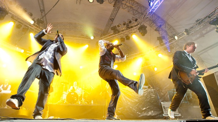 Madcon performen einen Song und werden von einem Gitarristen unterstützt. Beide Bandmitglieder haben ein Mikrofonin der Hand. Sie drehen sich jeweils von einander weg, beide haben einen Arm in der Luft