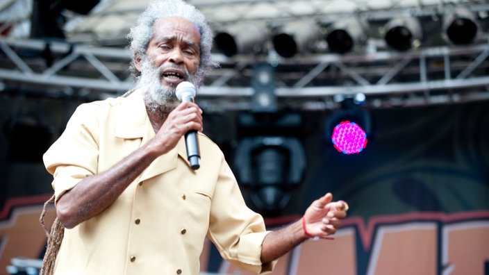 Reggae-Legende Max Romeo steht in gelben Hemd auf der Bühne. Er trägt einen grauen Rauschebart