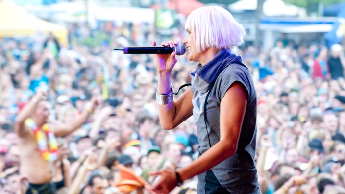 Sängerin Mono steht mit lila gefärbten Haaren auf der Bühne und singt. Unter ihr steht die zuschauende Menschenmenge
