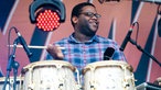 Der Drummer von Trombone Shorty schwingt die Sticks um Trombone Shorty zu unterstützen. Er blickt zur Seite