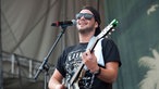 Mann mit Sonnenbrille und umgedrehter Kappe spielt Gitarre und singt in ein Mikrofon
