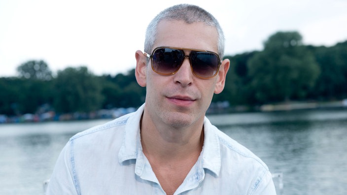 Mann mit kurzen grauen Haaren und großer Sonnenbrille am Seeufer