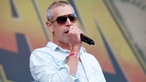 Mann mit kurzen grauen Haaren und großer Sonnenbrille singt in ein Mikrofon