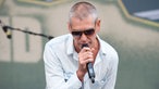 Mann mit kurzen grauen Haaren und Sonnenbrille singt in ein Mikro und schuat nach unten