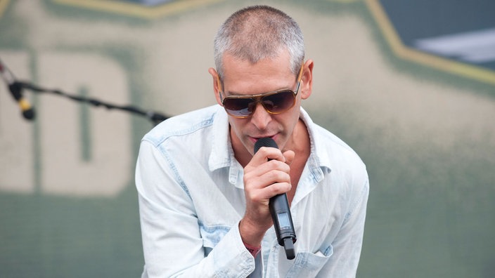 Mann mit kurzen grauen Haaren und Sonnenbrille singt in ein Mikro und schuat nach unten