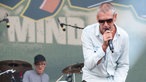 Mann mit kurzem grauen Haar und Sonnenbrille singt in ein Mikro, im Hintergrund ein Schlagzeuger